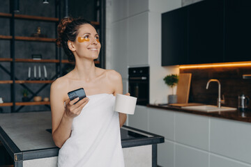 Happy woman with under eye patches holding smartphone and morning coffee got good news message