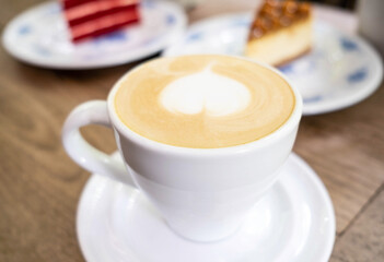 A white cup of latte art coffee with heart shape on old wooden background and there are two peices of cake behind the cup