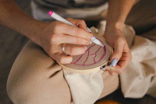 Close-up view of a woman's hands holding a pink marker and drawing an outline on the canvas she is going to embroider. Doing cross-stitch handwork. Closeup.