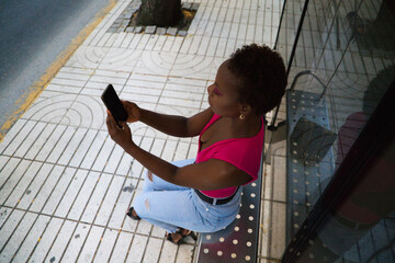 Young and beautiful Afro American woman sitting at the bus stop consulting her mobile phone and taking pictures of herself while waiting for the bus. Public transport concept.