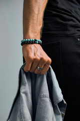 Cropped close-up shot of a man's hand with a ring and blue and black bracelets made out of volcanic and decorative stones. The man is holding a denim jacket on a gray background. Side view.