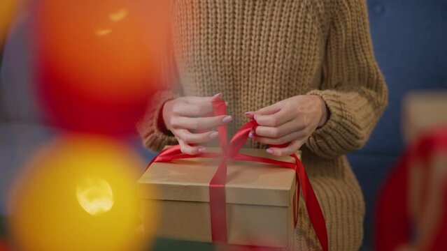 The girl unpacks a Christmas present. Close-up of a woman holding a Christmas gift box with a red ribbon in her hands. A woman in a sweater celebrates the New Year and opens a gift found under the tre