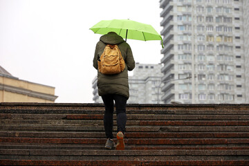 Woman with green umbrella walking up the steps on city buildings background. Rain in autumn city