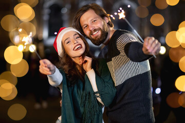 cheerful couple outdoors in winter waiting for the celebration with sparklers in their hands. The girl wears a Santa Claus hat and a man in a sweater