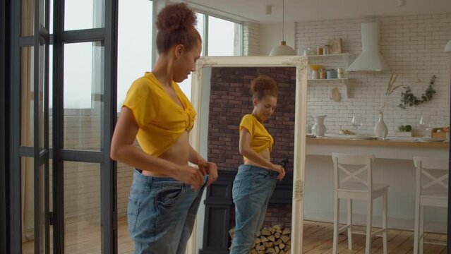 Cheerful charming black woman in too big jeans looking at reflection in mirror, demonstrating and enjoying weight loss diet achievement, expressing excitement and happiness in domestic kitchen.