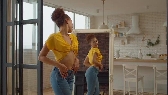 Portrait of joyful charming African female in trendy clothes looking at reflection in mirror, enjoying appearance and shapes, slapping on buttocks', expressing cheerful mood and excitement.