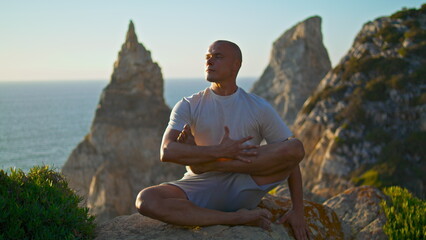 Naklejka na ściany i meble Handsome man performing yoga exercise in morning sunlight beautiful ocean rock.