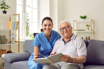 Fototapeta Senior man enjoying quiet pastime in retirement home. Portrait of nurse together with eledrly patient. Happy old male patient and young female nurse sitting on sofa and reading book together obraz