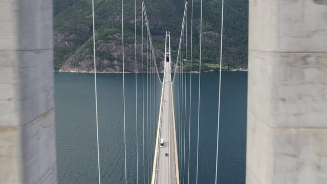 Several vehicles drive over the single-lane suspension Hardanger Bridge over the Eidfjord with a tunnel directly into the mountain in Noordwegen. Drone dolley shot between the pillars