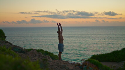 Sunset yogi silhouette training standing head at ocean cliff. Fit man practicing