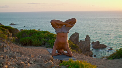 Muscular guy practicing yoga asana at beautiful sunset landscape on ocean cliff.