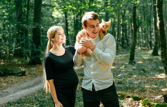 Young pregnant woman, her husband and their red cat in park. Happy family outdoors. Expecting for firstborn