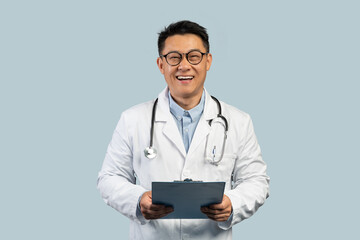 Cheerful middle aged korean man doctor in white coat and glasses with tablet, isolated on blue background