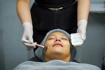 Closeup cosmetologist use cosmetic brush applying facial jelly's gel on woman client face in beauty clinic.