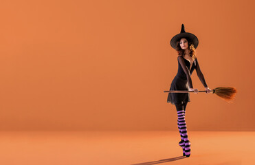 A ballerina on pointe shoes in a black witch costume in a hat and with a broom dances on an orange background.