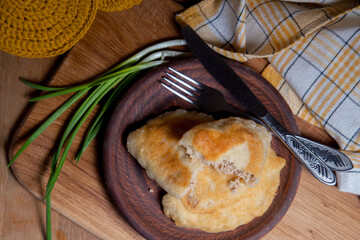 Clay plate of fried meat pies with cutlery and green onion on wooden table.