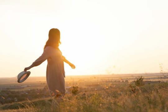 Back view of a cute girl with a dress and hat dancing in a field during golden hour