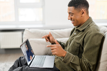 Side view of the concentrated male student involved virtual meeting, webinar. Black man making video call on the laptop, sitting on the sofa at home, talking and gesturing, while explaining something