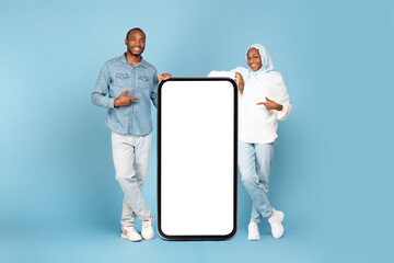 Black Muslim Couple Pointing At Huge Cellphone Screen, Blue Background