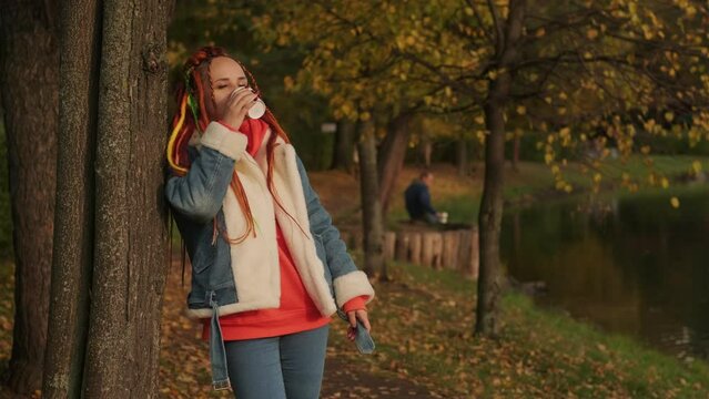 Young woman with dreadlocks in warm clothes standing near tree and drinking coffee. Female in casual outfit standing near green trees and drinks hot coffee or tea while resting in park on sunny day