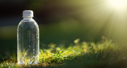 A plastic bottle with clean drinking water stands on the grass in the rays of the rising sun. Cool refreshing water in a bottle with dew drops .