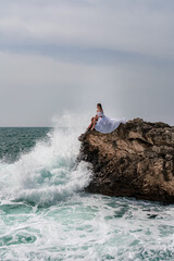 A woman in a storm sits on a stone in the sea. Dressed in a white long dress, waves crash against the rocks and white spray rises above her.