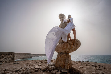 A woman stands on a stone sculpture made of large stones. She is dressed in a white long dress,...