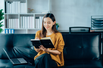 Fototapeta premium Portrait of a smiling Asian woman sitting on a sofa. Using laptop in the living room at home, working at home via online video conference.