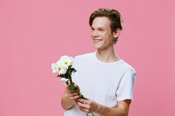 joyful, cute young man stit in a white T-shirt on a pink background with a bouquet of daisies in his hands