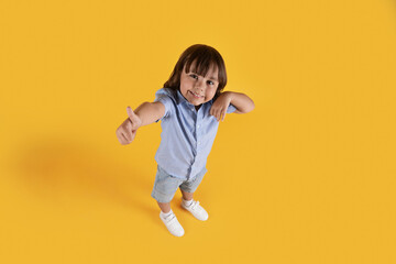 Above view portrait of little boy showing thumb up gesture, smiling to camera, orange studio background, full length
