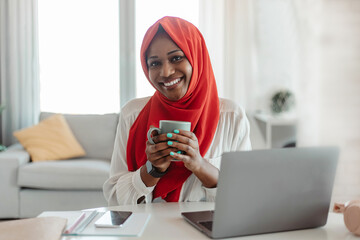Happy black islamic businesswoman working on laptop and drinking coffee while working at home office
