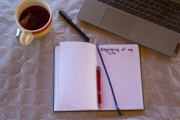 Image of an open diary with the heading Beginning of my life, with a blank page to insert text, a pen and a laptop.
