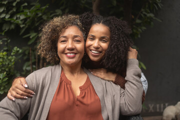 A young African-American woman and her mother are hugging, smiling, and posing on the patio in front of lush greenery.