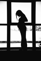 silhouette of a beautiful sexy pregnant brunette woman staying near windowand holding her belly