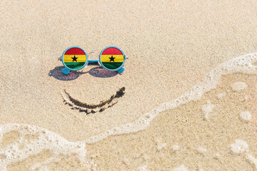 Fototapeta na wymiar A painted smile on the sand and sunglasses with the flag of the Ghana. The concept of a positive and successful holiday in the resort of the Ghana.