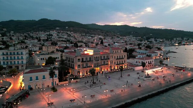 Flying over the main square of Spetses at coastline revealing the Poseidonion Grand Hotel, greec houses, pier, yachts and landscape as sunset twilight in the evening