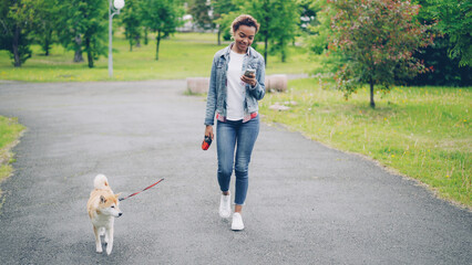Pretty African American girl in denim jacket and jeans is walking her purebred dog in city park and using smartphone going along path with green lawns and trees visible. - Powered by Adobe