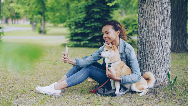 Attractive young African American girl is taking selfie with cute obedient dog resting in city park cuddling and caressing beautiful animal. Modern technology, loving pets and nature concept.