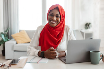 Portrait of black muslim freelancer woman in hijab sitting at table with laptop and smiling at...