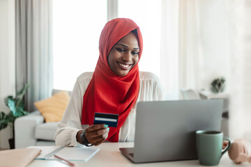 Cashless payment. Happy black muslim woman in hijab holding credit card and using laptop, shopping online from home