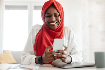 Mobile communication concept. Happy black islamic lady in headscarf using smartphone, enjoying...