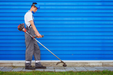 Young worker in overalls mows grass with trimer against background of blue metal siding. Janitor...
