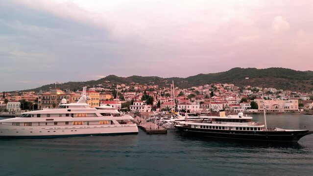 Flying backwards at the yacht pier in front of the Poseidonion Grand Hotel and main squar of Spetses revealing the cityscape and coast at sunset