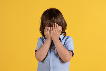 Kids offence. Close up portrait of unrecognizable little boy crying, covering face with hands, orange studio background