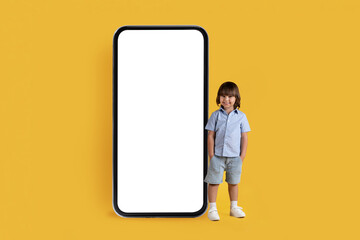 Adorable little boy with hands in pockets standing nera huge smartphone with blank white screen, empty space