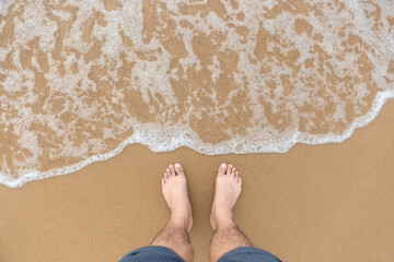 The feet of man without any shoe standing on the smooth beach and softly foam from wave of the sea by top view photography. Copy space of sea and beach of photo