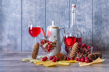 On wooden table there is bottle and glasses filled with red wine, coffee pot and mugs for drink, vase with ripe cherries, yellow maple leaves and fir cones. - Powered by Adobe