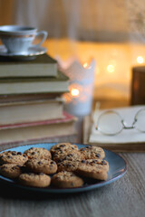 Plate of chocolate chip cookies, stack of vintage books, reading glasses, cup of tea or coffee, lit candle and fairy lights. Hygge at home. Selective focus.