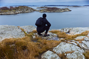 Obraz premium Hiker in black gear with a backpack crouching on a rocky cliff, looking out over a calm sea and scenic archipelago islands under a moody, overcast sky. Perfect for nature and adventure concepts.