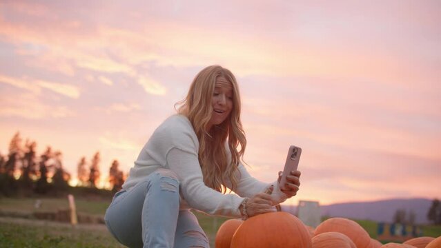 Low angle of female content creator takes selfie video at pumpkin patch during sunset